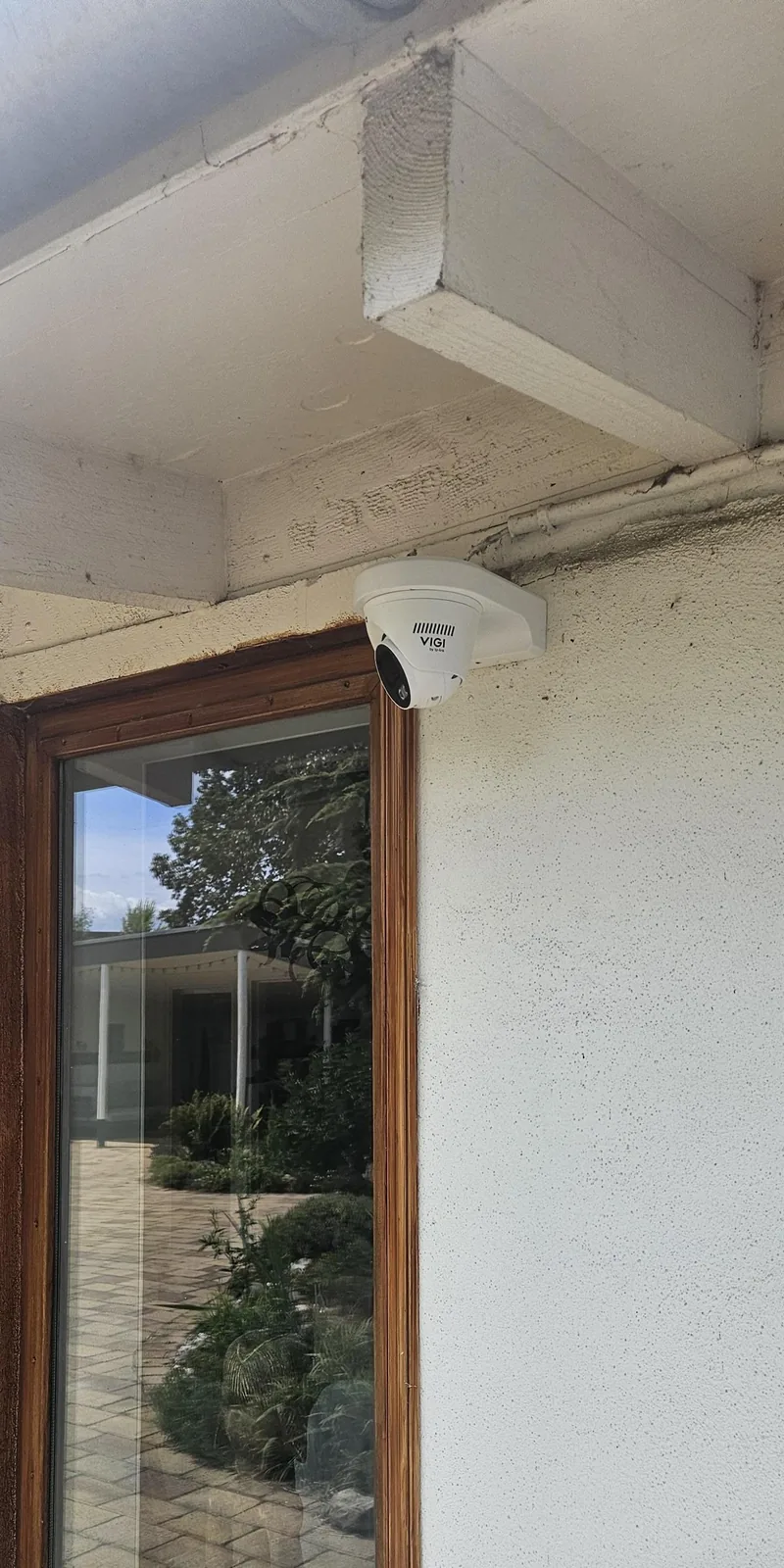 Witte VIGI-turretcamera in de hoek boven een houten raamkozijn, onder een overstek met witte balken