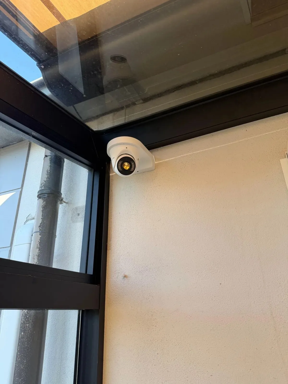 Witte VIGI-turretcamera in de hoek onder een glazen overkapping, naast een zwart raamkozijn