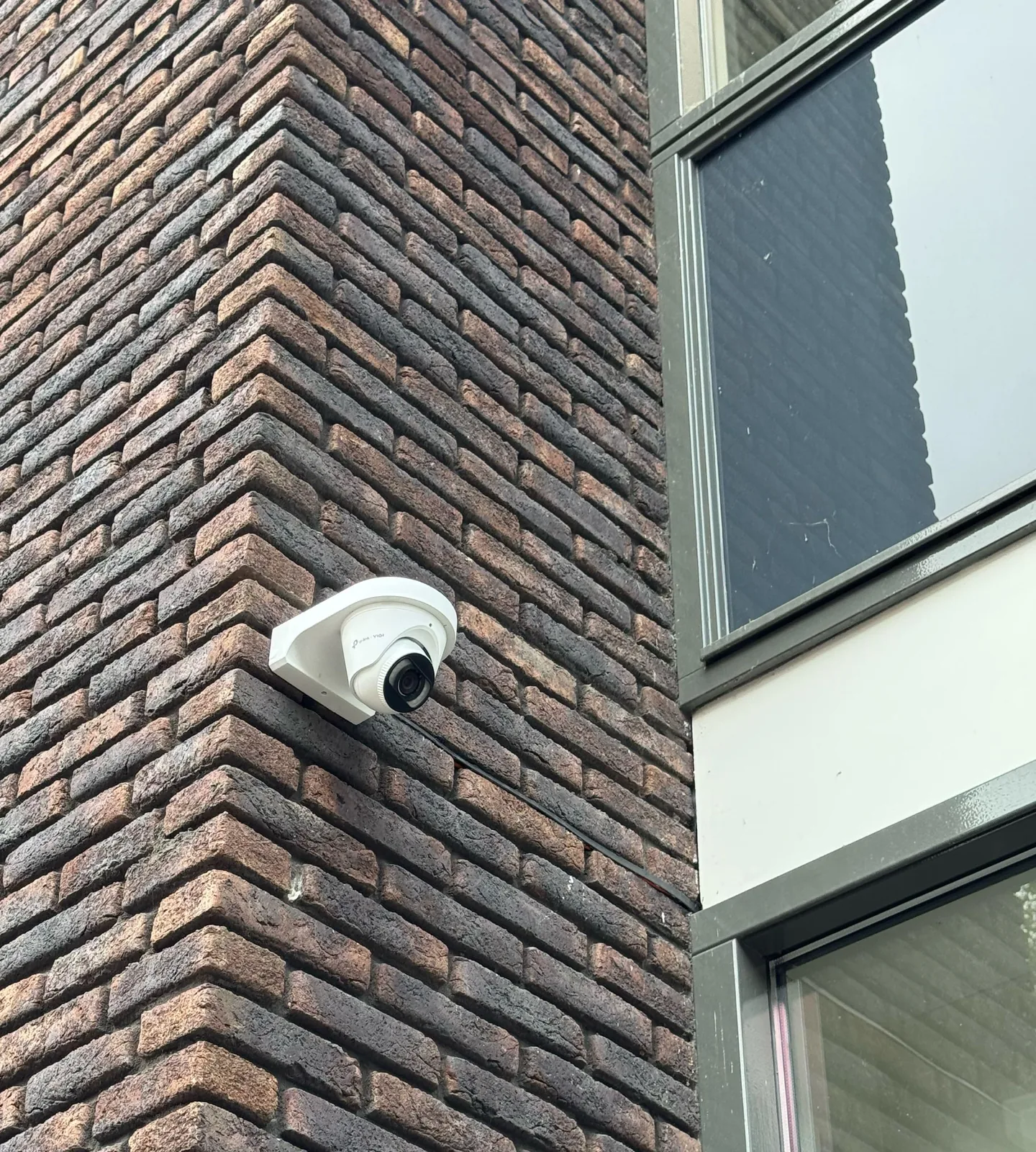 Witte VIGI-turretcamera op een muurbeugel op de hoek van een donkere bakstenen gevel, naast een raam